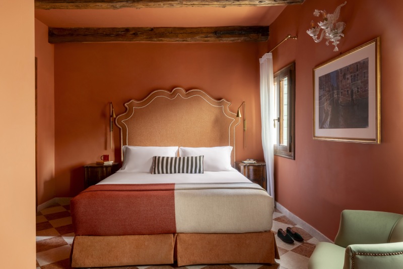 Queen bed with stylish upholstered headboard, striped accent pillow, and checkered tile floor in an orange room - Hotel Riva, BZAR hotels in Venice